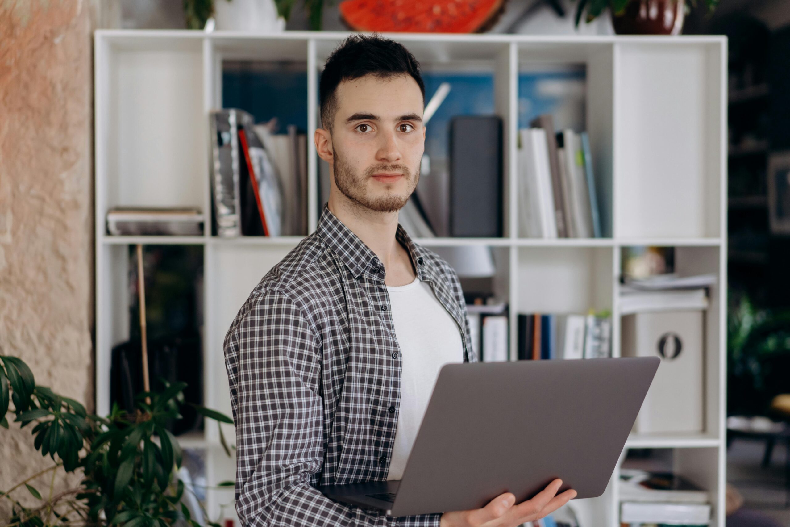 Man holding laptop in modern office setting with shelves. Professional and determined expression.